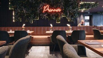 A modern restaurant with stylish seating areas and an inviting atmosphere. In the background, a neon sign reads "Prenner" against a green plant decoration.