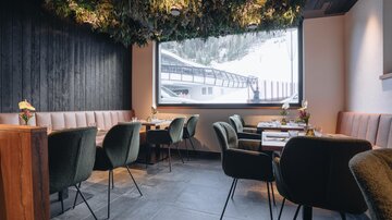 A modern restaurant with elegant green chairs and a cozy atmosphere. Through the large window, one can see a snowy landscape.