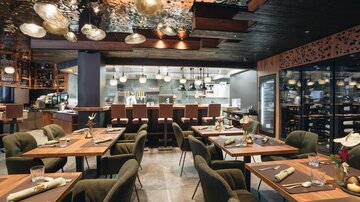 An elegant restaurant with modern furniture and warm lighting. The tables are beautifully set and an open kitchen is visible in the background.