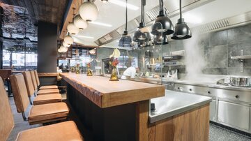 A modern restaurant with an open kitchen and a long wooden bar. The decor is stylish and the lighting creates an inviting atmosphere.