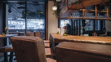 A stylish bar with wooden interior and modern seating. Large windows provide plenty of daylight and a welcoming atmosphere.