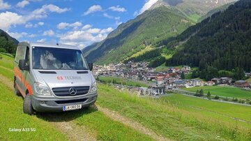 A van is standing on a meadow with a view of a picturesque village nestled in the mountains. The sky is blue with some clouds.