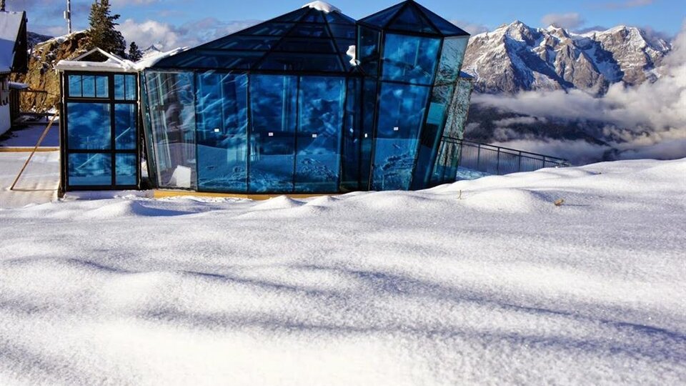 A modern glass building stands amidst a snow-covered landscape. In the background, impressive mountain peaks and a clear sky are visible.