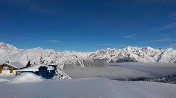 A winter landscape with snow-covered mountains and a clear blue sky. Traditional huts are visible in the foreground.