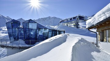 A modern glass building in a snow-covered mountainous landscape. Bright sun and snow-covered hills create a winter atmosphere.