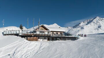 A mountain cabin with a terrace in the midst of a snowy landscape. In the background, snow-covered mountains and a clear blue sky can be seen.