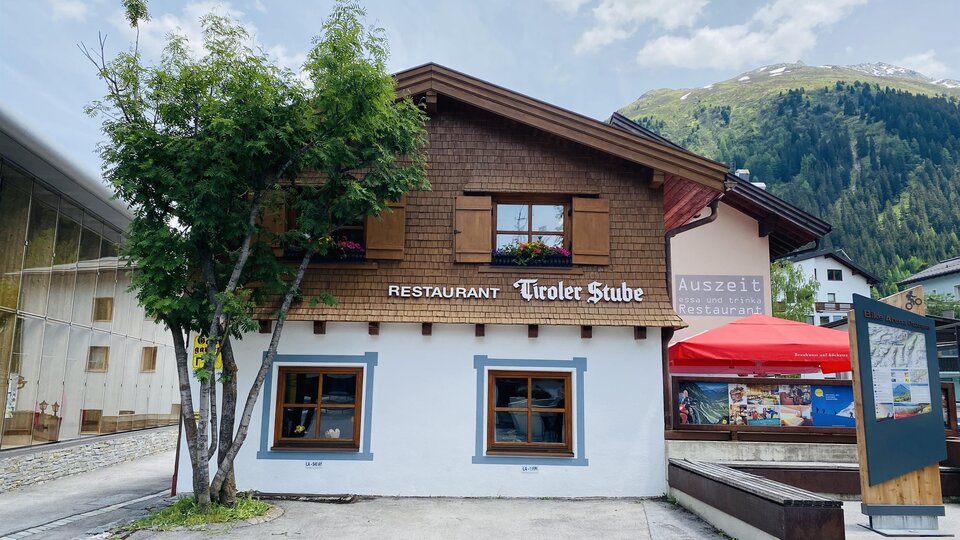 Ein gemütliches Restaurant namens "Tiroler Stube" mit Holzverkleidung und Fenstern. Im Hintergrund sind die Berge zu sehen.