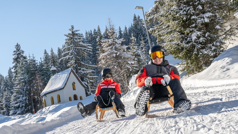 A snowy landscape with two people riding sleds. In the background, there are winter trees and a small, cozy house.