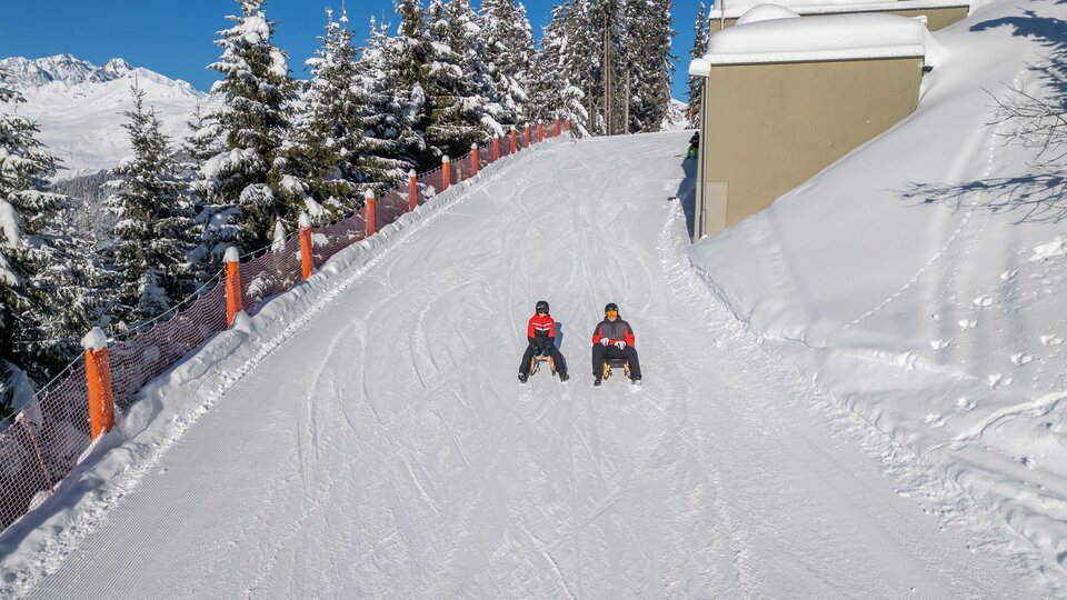 Two people are sitting on a snowy slope. Surrounded by snow-covered trees and a clear blue sky.
