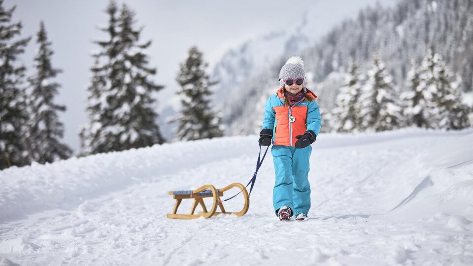 A child is pulling a sled along a snowy path. In the background, snow-covered trees and mountains can be seen. | © TVB Paznaun - Ischgl