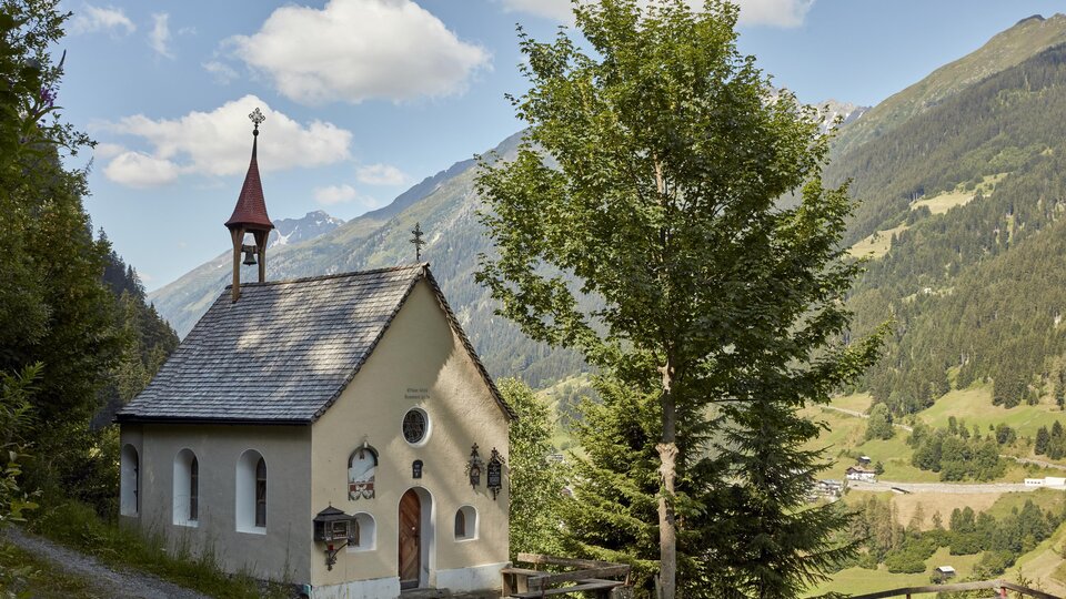 Eine kleine Kapelle umgeben von Bäumen und Bergen. Der Himmel ist partly cloudy und die Landschaft ist grün und malerisch.