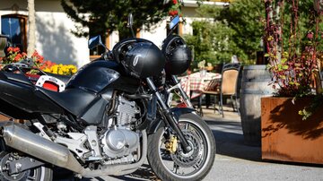 A black motorcycle is standing in front of a cozy restaurant with flowers and outdoor tables. In the background, green trees and a beautifully designed outdoor area are visible.