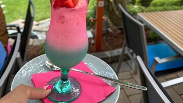 A colorful cocktail with strawberries and a waffle in a cozy outdoor area. In the background, a green garden landscape is visible.