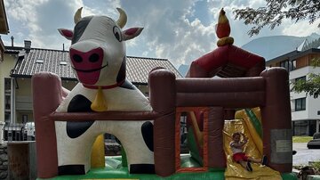 An inflatable cow with a sliding area is standing outside. The sky is cloudy and buildings are visible in the background.