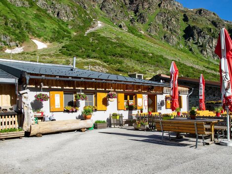 A cozy alpine hut with colorful flowers and red umbrellas. In the background, green mountains can be seen.
