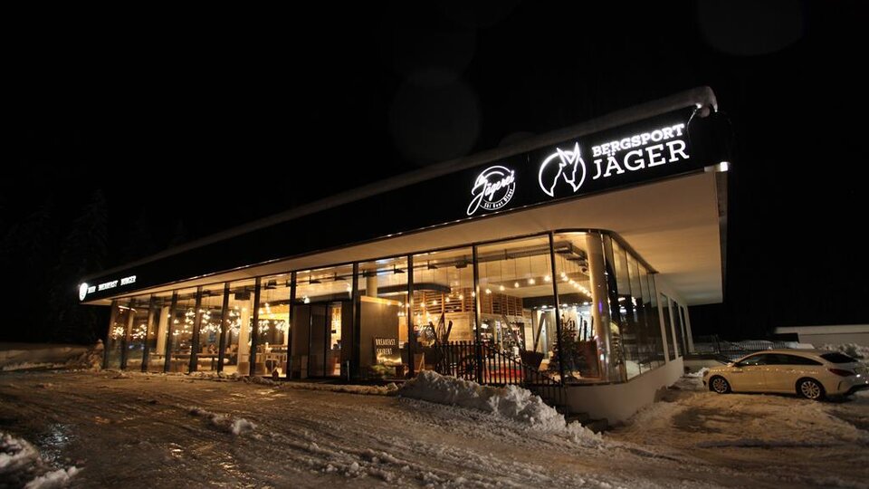 Ein modernes Sportgeschäft mit großen Fenstern, gut beleuchtet in der Nacht. Es ist von Schnee umgeben und der Geschäftsname "Bergsport Jager" ist gut sichtbar.