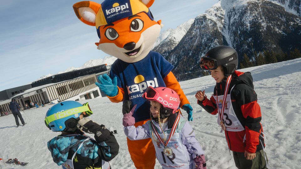 A mascot in the form of a fox stands on a snowy slope and interacts with three children. The children are wearing ski gear and are laughing.