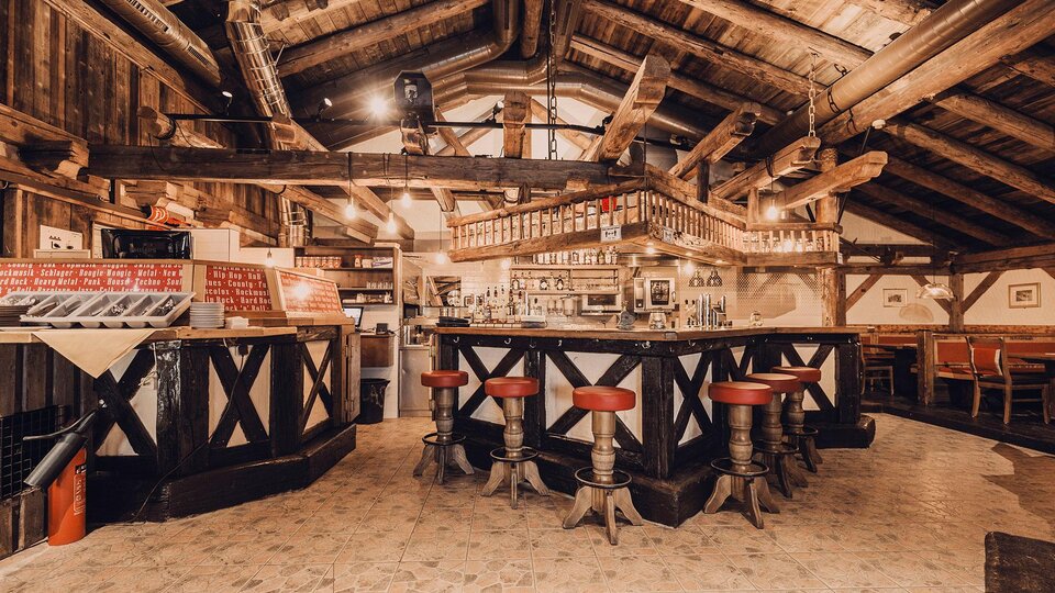 A cozy bar in a rustic ambiance with wooden ceilings and beams. Red bar stools surround the central counter.