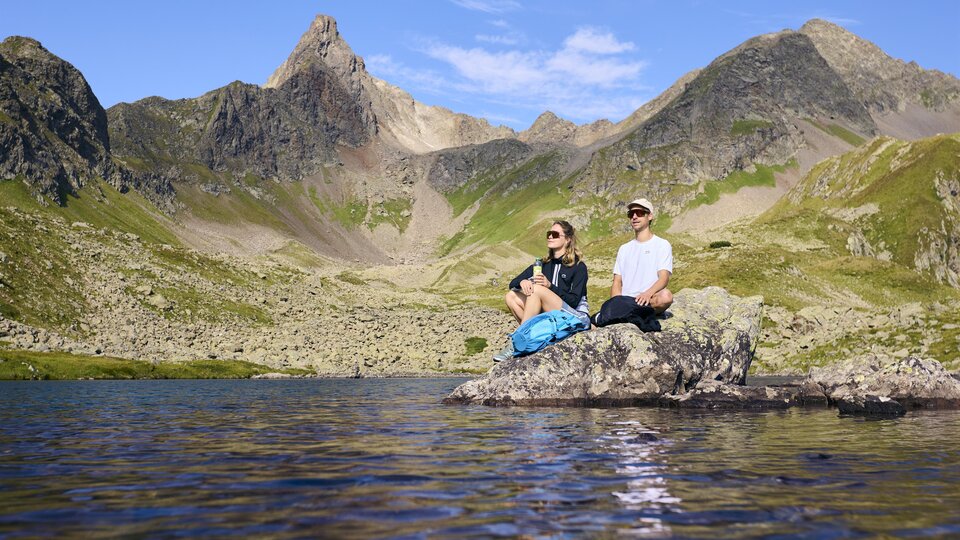 Two people are sitting on a rock at the edge of a lake in the mountains. The landscape is majestic with high peaks and a clear sky.