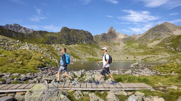 Two hikers are walking across a wooden path in a picturesque mountain landscape. In the background, green meadows and tall mountains can be seen.