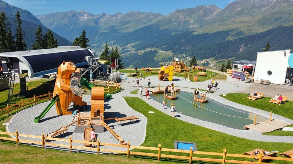Ein Spielplatz in den Bergen mit verschiedenen Holzspielgeräten für Kinder. Im Hintergrund sind grüne Wiesen und hohe Berge zu sehen.
