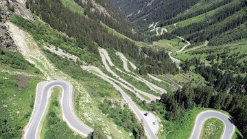 Eine kurvenreiche Straße führt durch eine grüne Berglandschaft. Die Berge und das Tal sind mit Bäumen bedeckt und der Himmel ist klar.