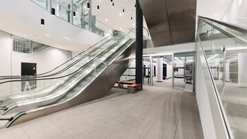 A modern lobby with bright walls and large windows. In the foreground, there are rolling stairs and a stylish seating area.