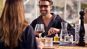 A man is sitting smiling at a table and enjoying a meal. In the background, glasses and a vinegar-oil set can be seen while he is talking to a person.