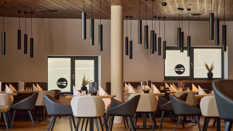 A modern restaurant with elegant tables and chairs. The ceiling is decorated with simple pendant lights and large windows provide plenty of light.