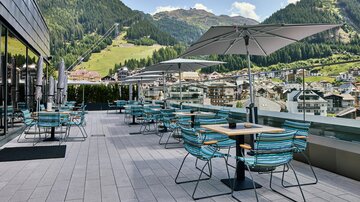 A cozy terrace with blue chairs and tables, surrounded by mountains. Umbrellas provide shade and the view of the city is breathtaking.