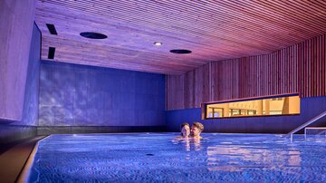 A modern swimming pool with soft lighting and wooden paneling. Two people enjoy the relaxing atmosphere in the water.