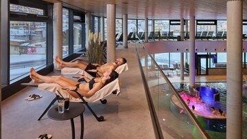 A relaxing wellness area with two people on loungers. In the background, there is a pool with colored lights and modern architecture.
