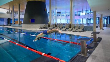 A modern indoor pool with a swimmer jumping into the water. The surroundings are bright and inviting, with loungers and large windows.