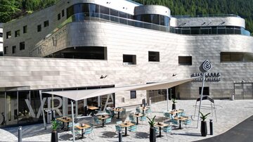 A modern building with an inviting terrace and tables. Green mountains are visible in the background.