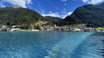 A picturesque landscape with a clear blue water basin and mountains in the background. The sky is blue with some white clouds.