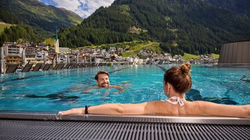 A beautiful infinity pool with a view of the mountains and a village in the valley. Two people enjoy the water and the breathtaking scenery.