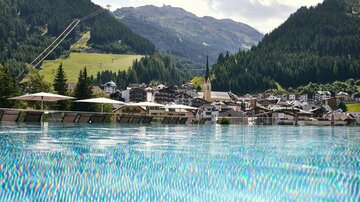 A beautiful view of a mountain village with green hills in the background. In the foreground, there is sparkling blue infinity pool water.