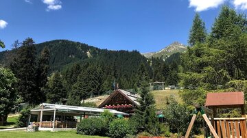 Eine schöne Landschaft mit Bergen und dichten Wäldern. Im Vordergrund befindet sich ein Gartencafé und ein Spielplatz.