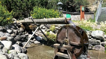 Ein Wasserrad, das von fließendem Wasser angetrieben wird, steht neben einem Fluss. Im Hintergrund sind grüne Pflanzen und ein Holzhaus zu sehen.