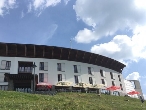 A modern building in the mountains, surrounded by meadows and blooming plants. The sky is clear and blue.
