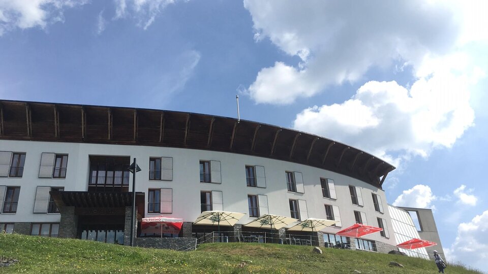 A modern building in the mountains, surrounded by meadows and blooming plants. The sky is clear and blue.