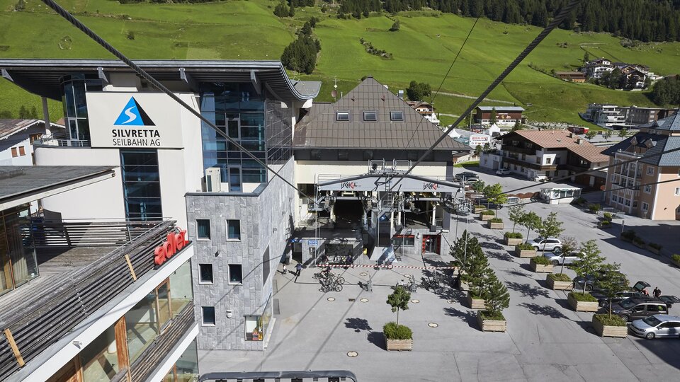 Ein moderner Liftbereich mit einem großen Gebäude im Hintergrund. Die Umgebung ist grün und bietet eine gemütliche Atmosphäre.