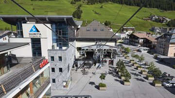 Ein moderner Liftbereich mit einem großen Gebäude im Hintergrund. Die Umgebung ist grün und bietet eine gemütliche Atmosphäre.