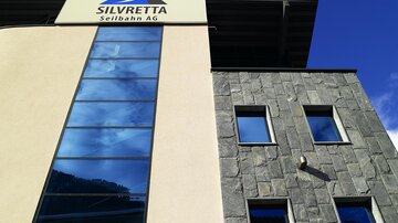 Ein modernes Gebäude mit einer Glasfassade und einem großen Schild der Firma Silvretta Seilbahn AG. Der Himmel ist klar und blau, was eine angenehme Atmosphäre schafft.