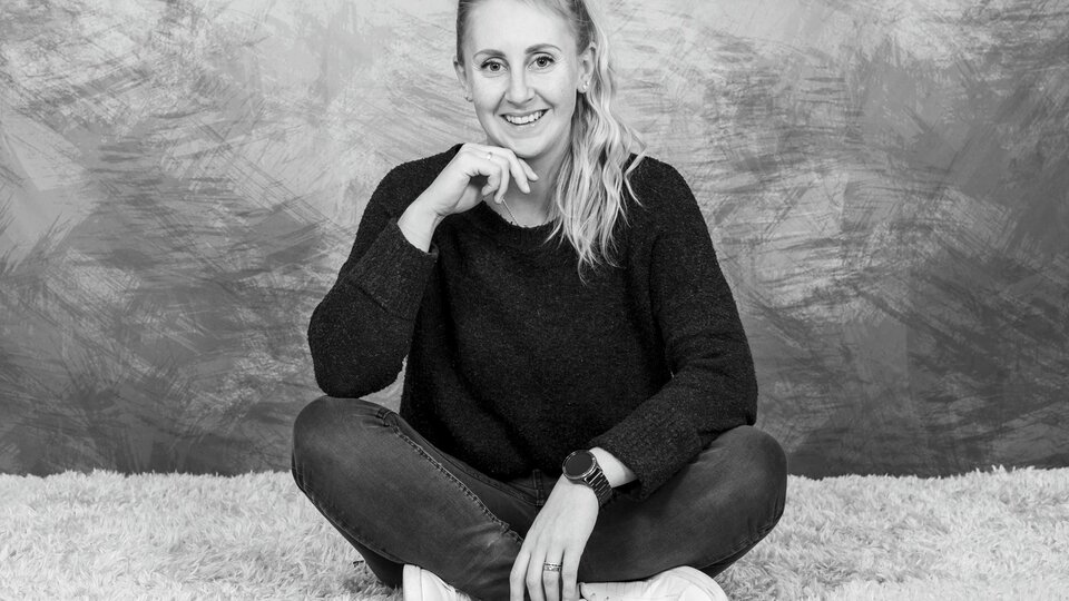 Eine junge Frau sitzt lächelnd mit gekreuzten Beinen auf einem weichen Teppich. Sie trägt einen dunklen Pullover und Jeans. | © Simone Drexel Photography
