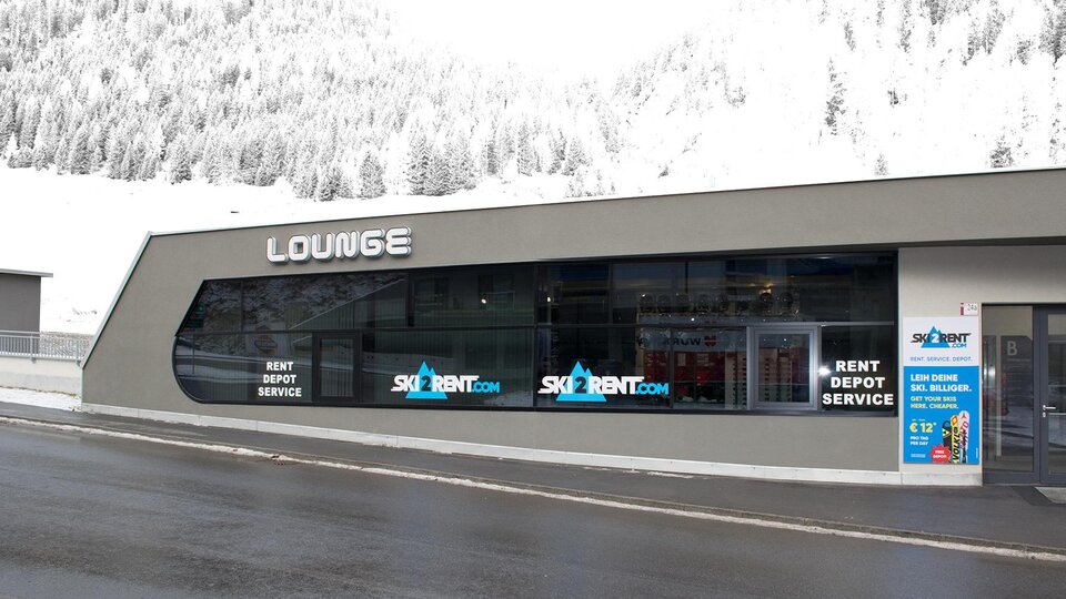 A modern ski rental shop with large windows. It is located in a snow-covered environment and offers a cozy lounge area.