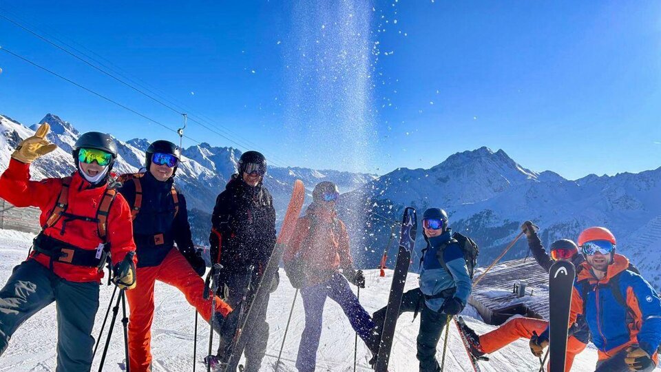 A group of six skiers stands in the mountains. The sky is clear and the sun is shining, as snow drifts down on the group.