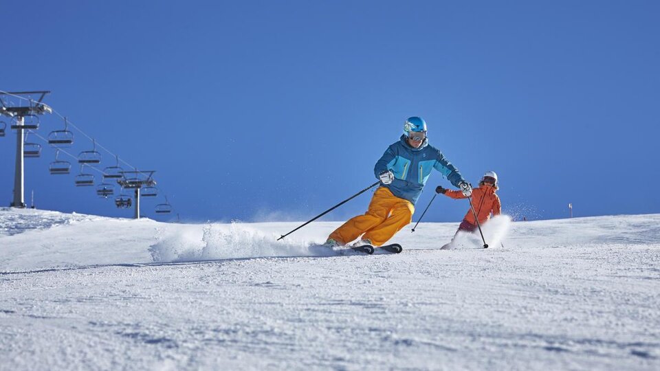 A skier is dynamically descending the slope while a child in orange ski clothing follows him. The sky is bright blue and the slope is covered with fresh snow.