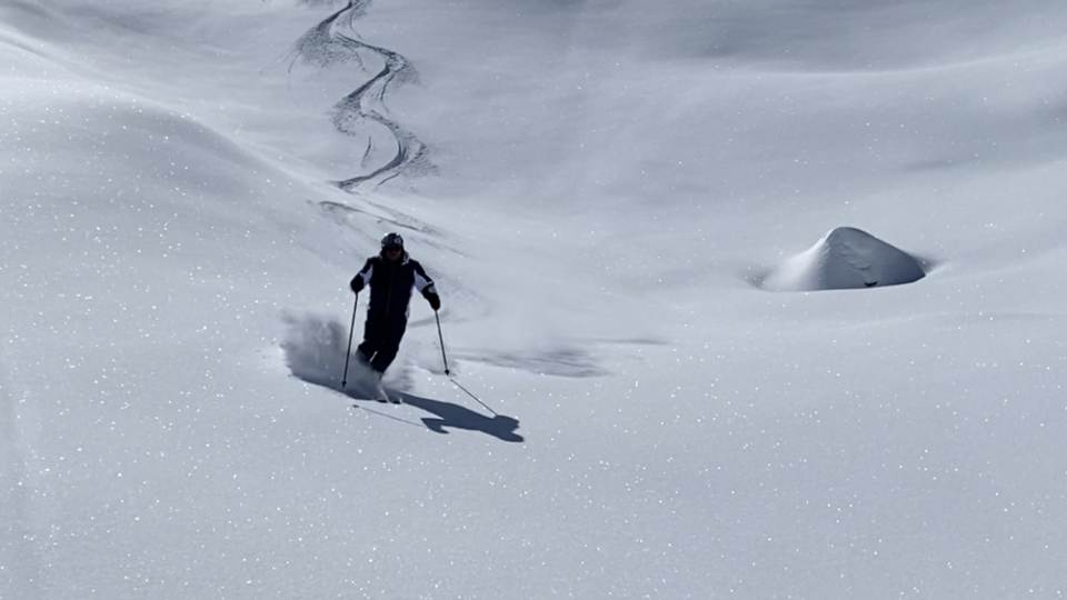 Ein Skifahrer geht durch tiefen, glitzernden Schnee unter einem klaren blauen Himmel. Im Hintergrund sind sanfte, verschneite Hügel zu sehen.