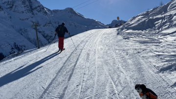Ein Skifahrer fährt eine schneebedeckte Piste entlang, während ein Hund neben ihm sitzt. Die Sonne scheint klar am blauen Himmel.
